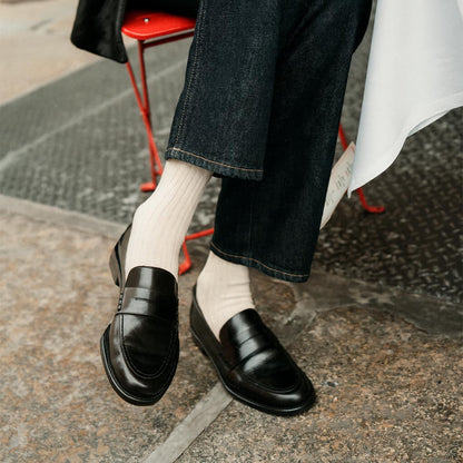 A person in dark denim pants, white ribbed socks, and margaux-development’s The Marlowe Loafer in Black Calf sits outdoors on a red metal chair with legs crossed—a timeless style staple.
