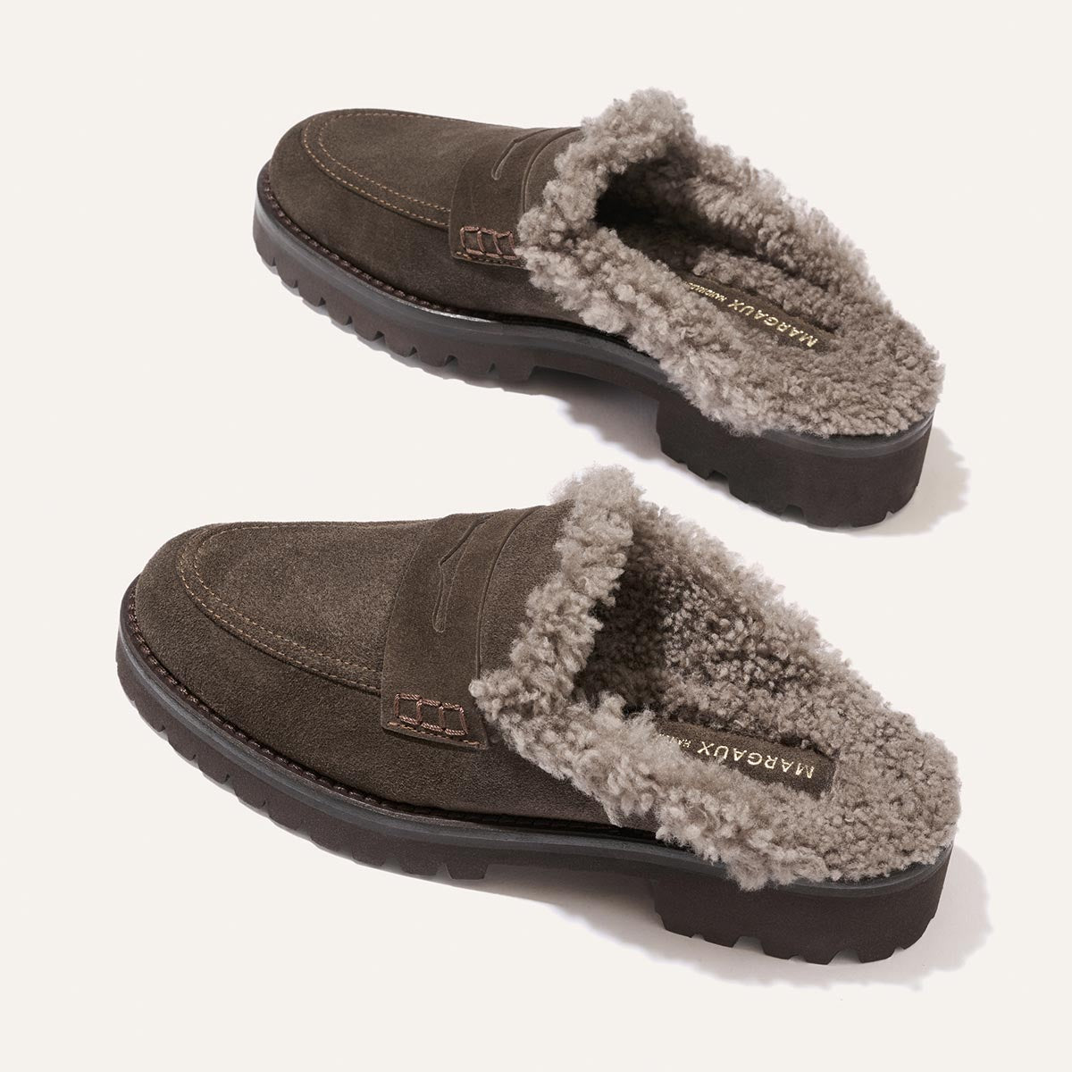 A pair of The Marlowe Mule in English Brown Suede and Shearling by Margaux, featuring rugged black soles and plush shearling lining, are displayed on a light background for a cozy feel.
