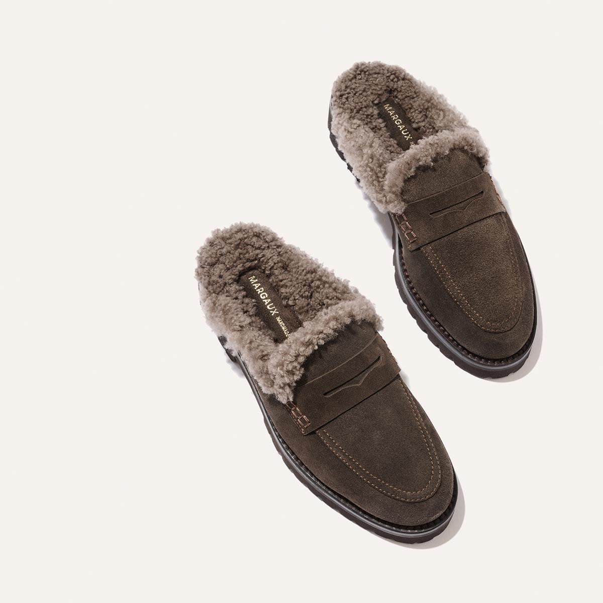 A pair of The Marlowe Mule loafers by Margaux in English Brown Suede with a cozy shearling lining are shown on a plain white background, featuring “MADE OF JAK” on the insole.