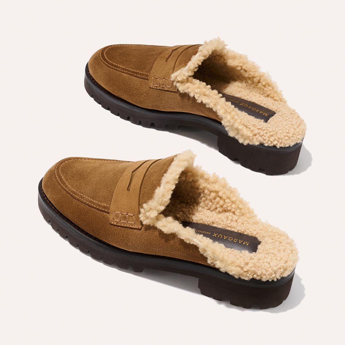 A pair of Margaux’s The Marlowe Mule in walnut suede with plush cream shearling lining and dark rubber soles is displayed diagonally on a light background.