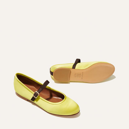 The Demi Jane - Citron Satin flats by Margaux feature elegant yellow satin with brown straps and buckles. One sits upright, the other displays its tan sole, both on a clean, pale background.
