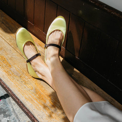 A person sits on a wooden bench with legs crossed, embodying elegant femininity in Margaux’s The Demi Jane - Citron Satin shoes. Sunlight highlights the light yellow satin finish and sophisticated dark brown straps.