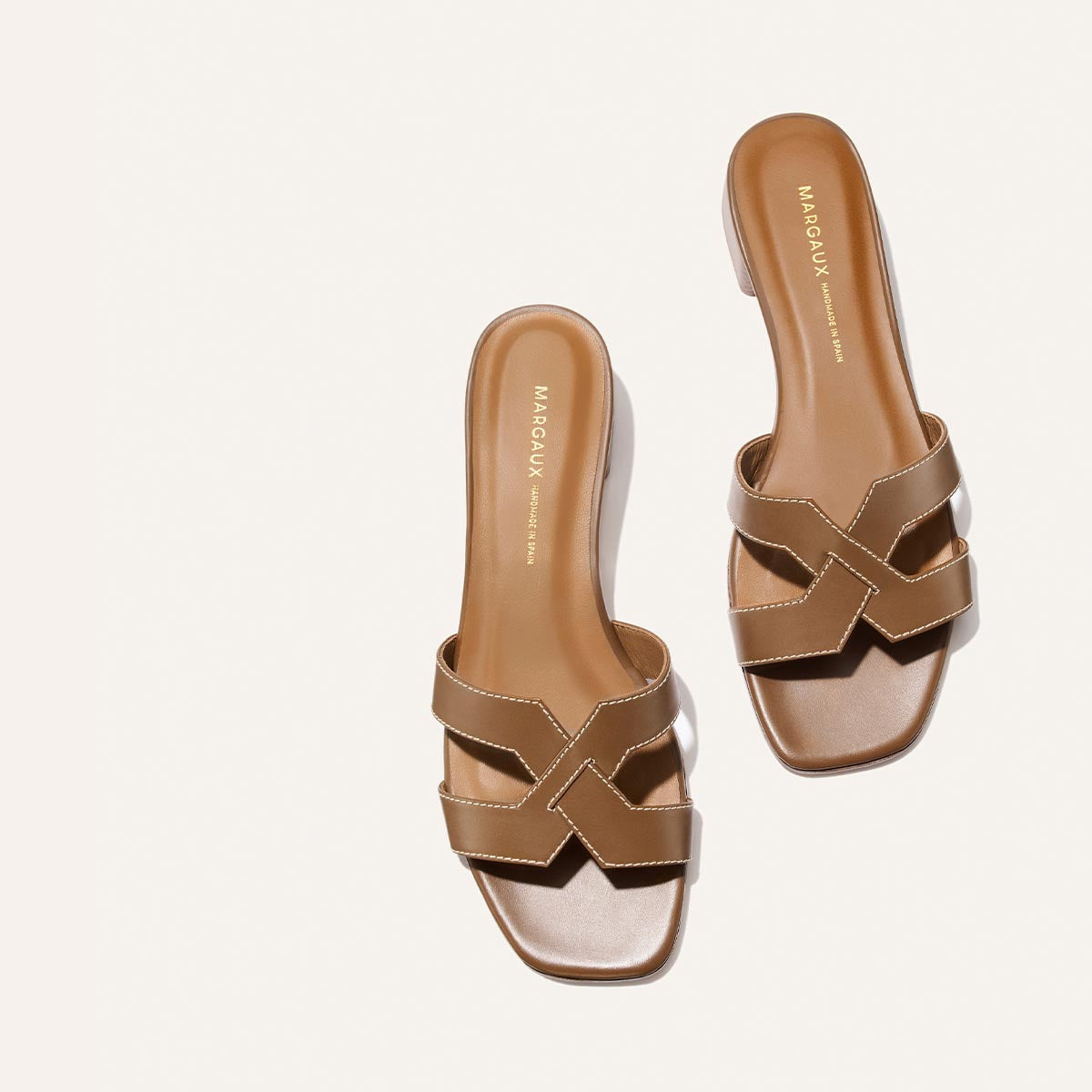 The MX 35 - Mocha Calf by Margaux features tan slide sandals with open toes, crisscross straps, white stitching, and the Margaux name in gold on the insole—all displayed on a plain white background for a chic, walkable look.