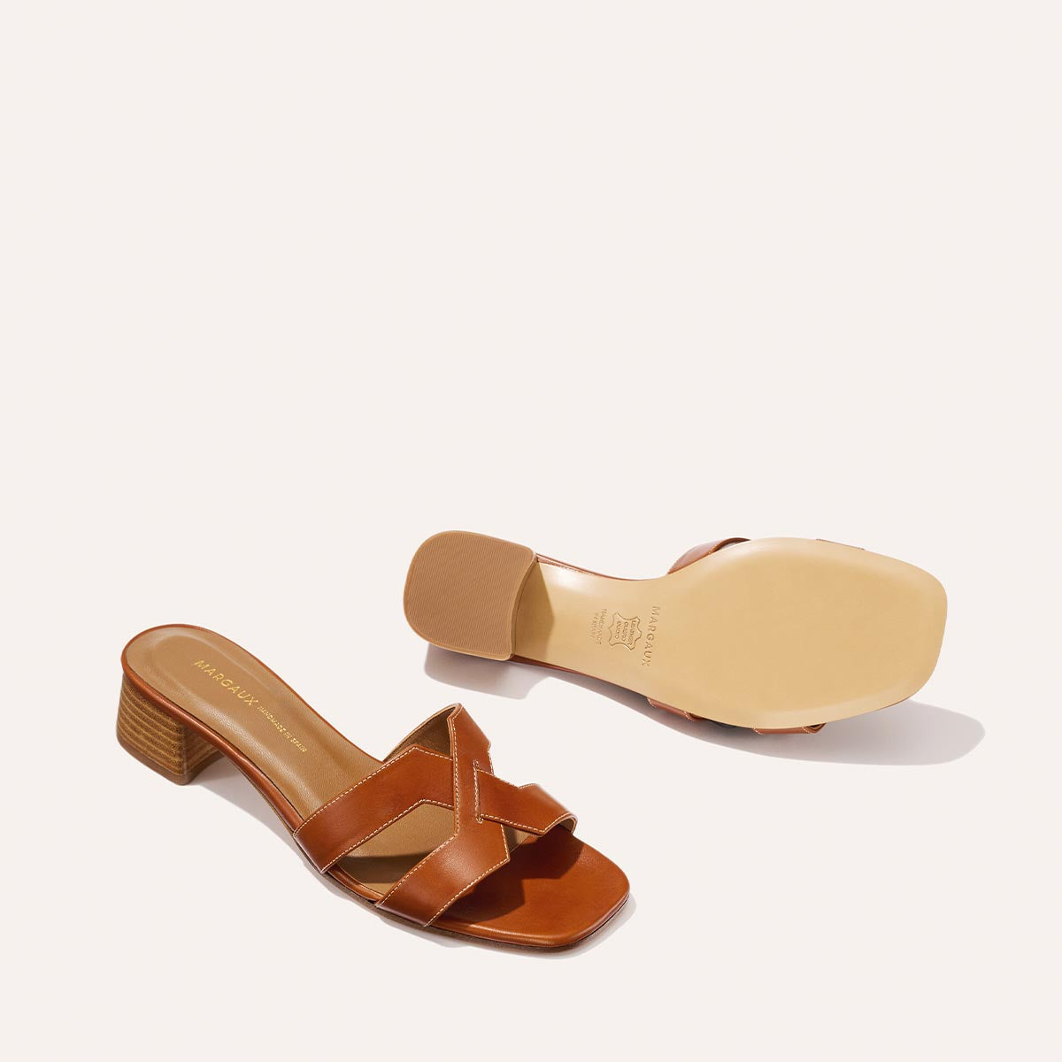 A pair of Margaux-development’s The MX 35 - Saddle Calf block-heeled sandals with crisscross leather straps is shown: one upright revealing the top and heel, the other on its side to display the sole, against a plain light background.