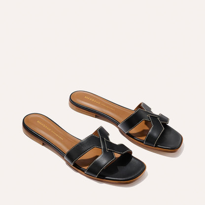 The MX Sandal - Black Calf by Margaux features black leather slides with open toes, brown insoles, low heels, stylish cut-out details, and white edge stitching.