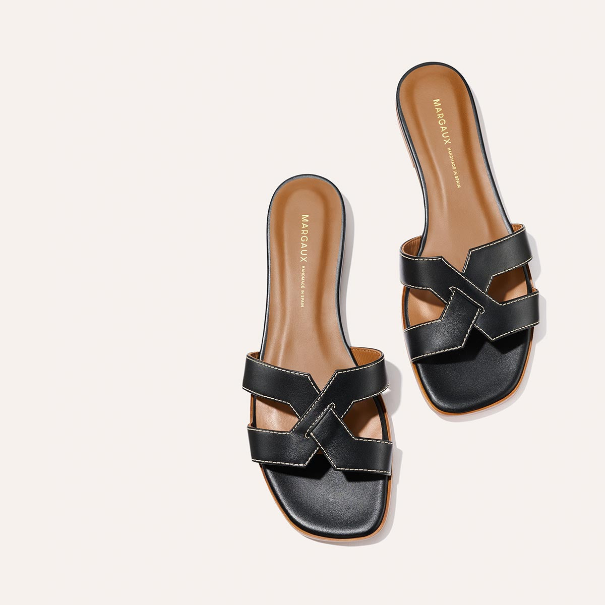 The MX Sandal - Black Calf by Margaux features black leather slides with tan insoles and the Margaux logo in gold on the heel, shown against a light beige background.