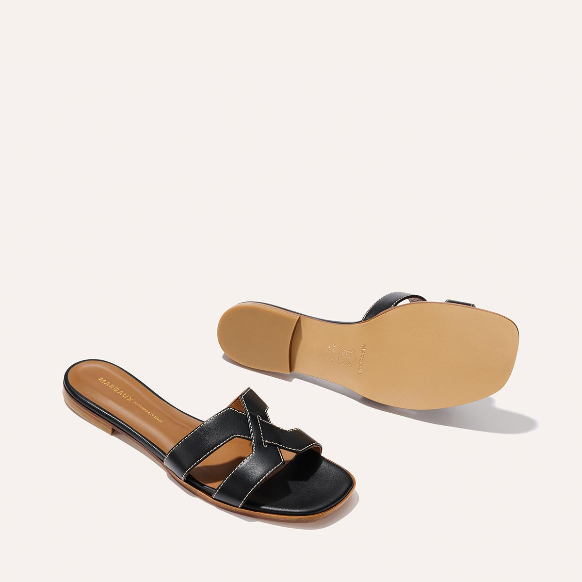 Two Margaux The MX Sandal - Black Calf slip-ons are shown: one displays the crisscross leather top, while the other is flipped to reveal its tan sole.