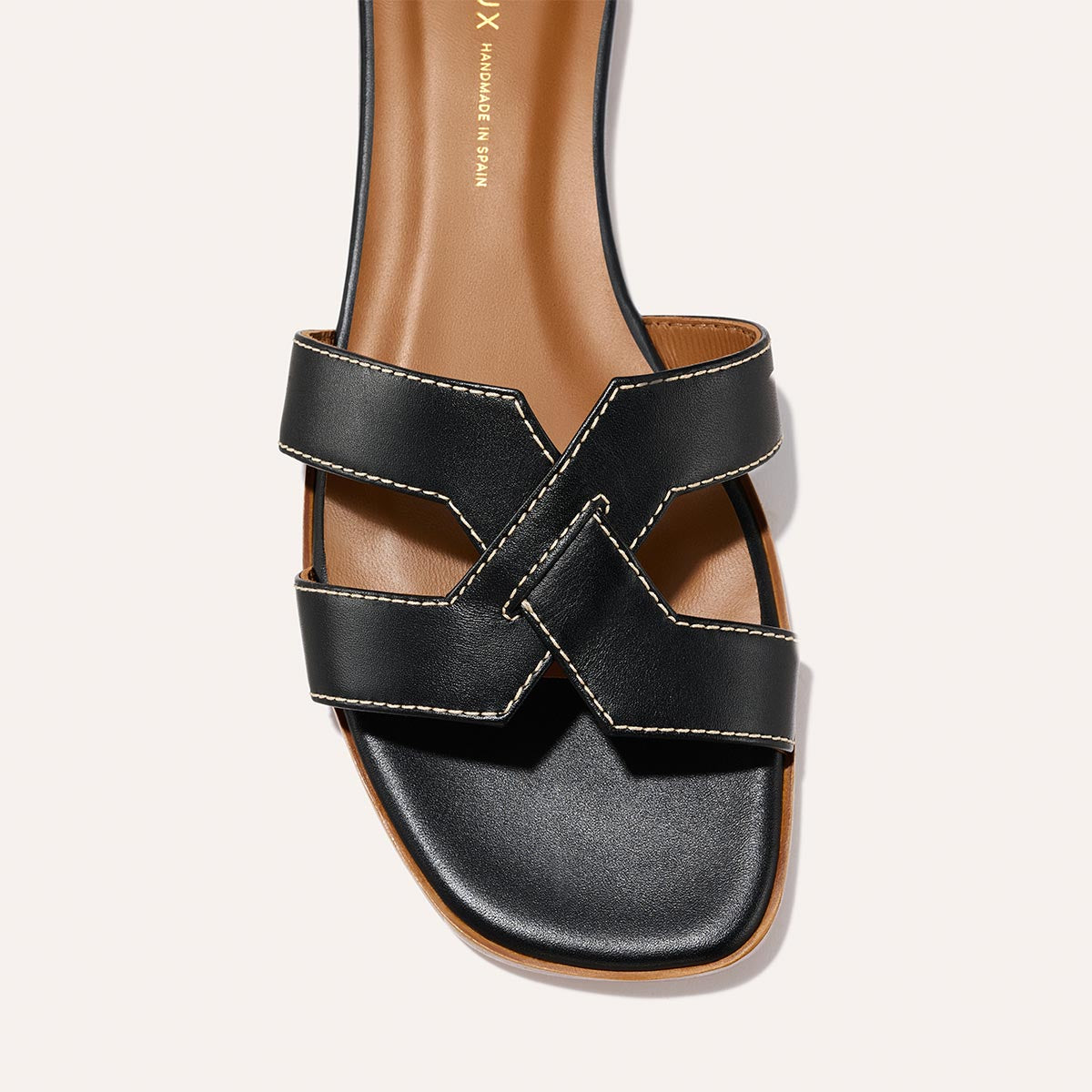 A close-up of Margaux’s The MX Sandal - Black Calf, showcasing two wide crisscrossed black leather straps with white stitching, a square toe, and a tan insole.