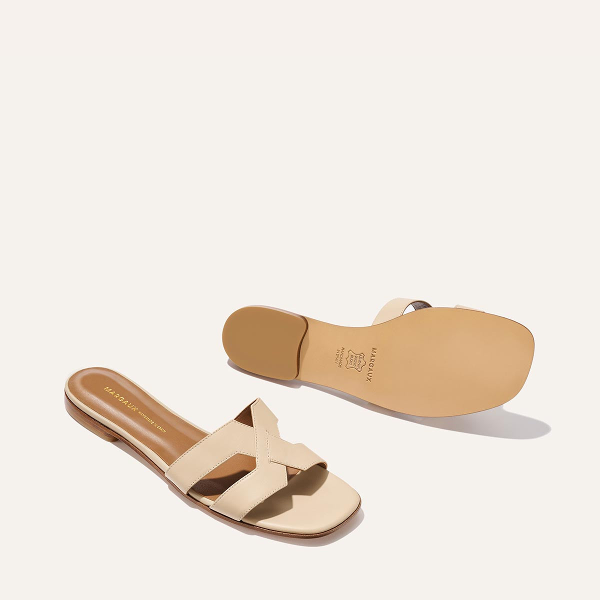 The MX Sandal - Latte Calf by Margaux: luxe beige leather slides with crisscross straps and flat soles—one showing the cushioned footbed, the other the sole—ideal for elevating daily style, photographed on a light background.