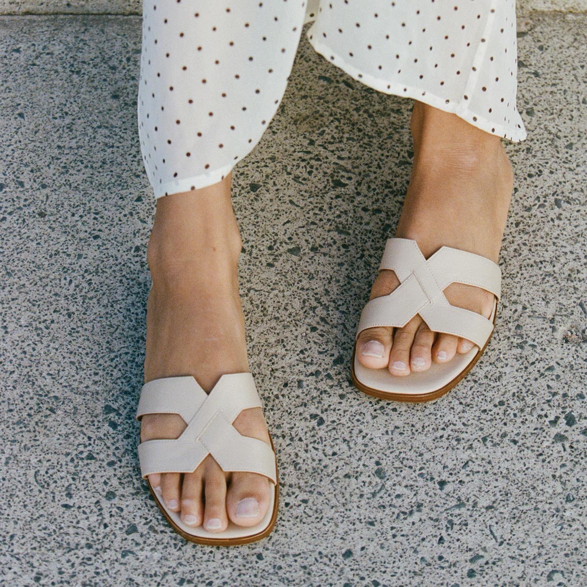 A person in white pants with black polka dots stands on a textured concrete surface, wearing Margaux’s The MX 35 - Latte Calf block heels with crisscross straps.