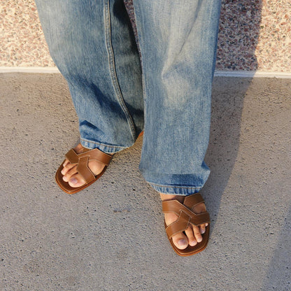 A person in loose blue jeans and walkable Margaux The MX 35 - Mocha Calf block heel sandals stands on a concrete sidewalk near a textured wall.