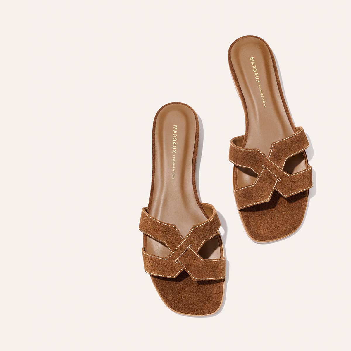 The MX Sandal - Saddle Suede by Margaux features brown suede crisscross straps, flat soles, and luxe leather craftsmanship. Photographed on a light background, the insole showcases Margaux branding.