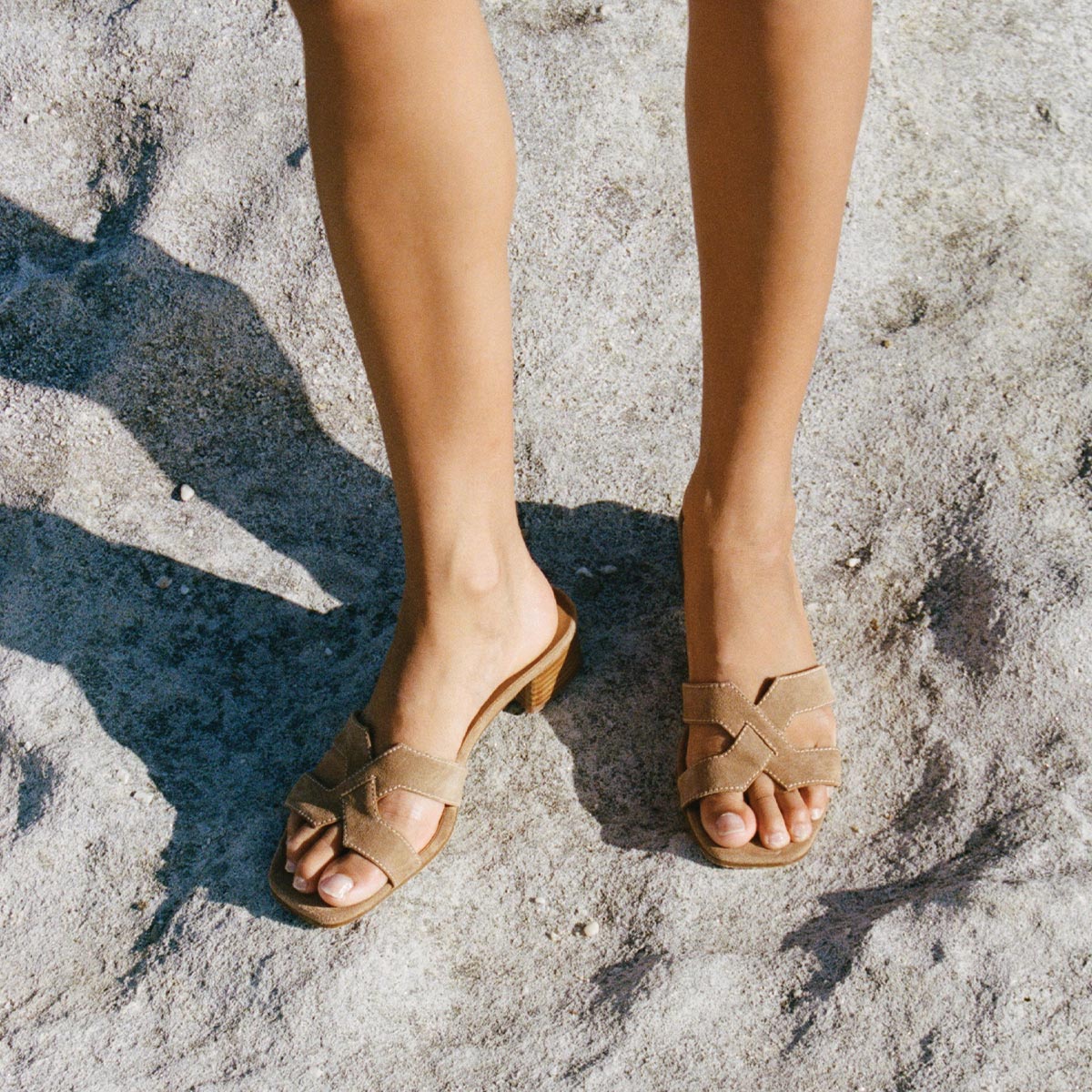 A person stands on sandy ground wearing Margaux’s The MX 35 - Biscotti Suede sandals; only their lower legs and feet are visible.