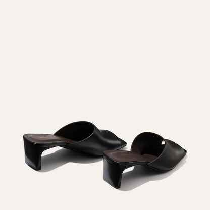 Margaux's The Naomi 50 in Black Nappa: black open-toe slip-on heeled sandals with wide crossed leather straps and chunky square block heels, displayed on a light beige background.