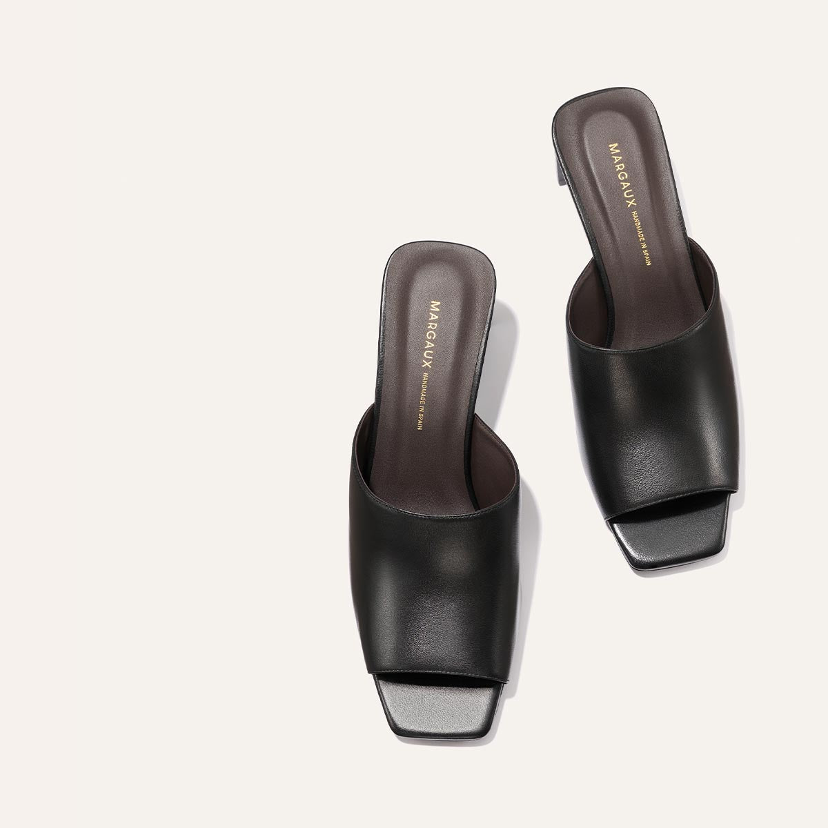The Naomi 50 - Black Nappa by Margaux: black leather open-toe slide sandals feature a square toe, low square block heel, and soft leather upper with dark brown insoles stamped in gold with Margaux branding, shown on a light background.