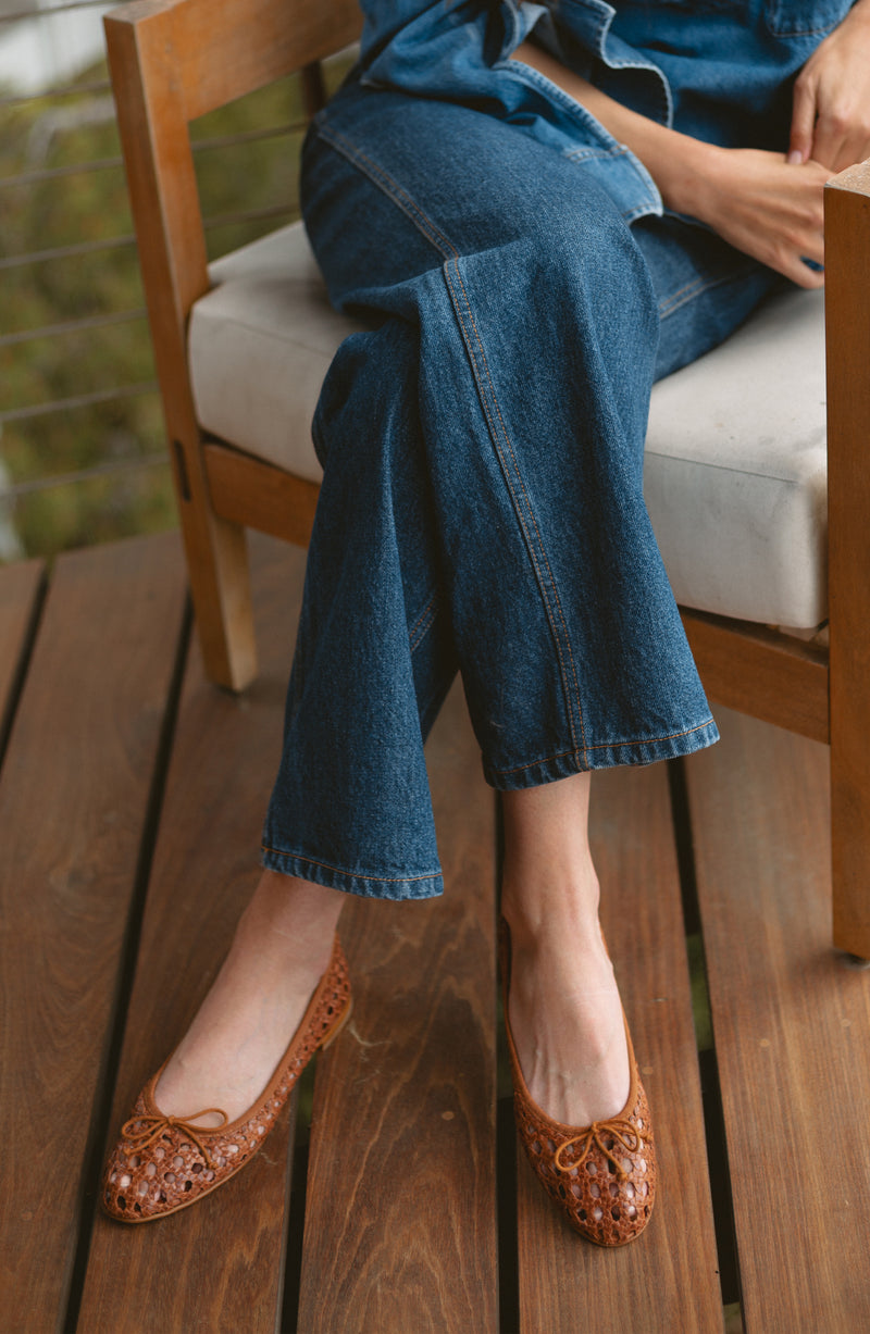 A person sits on a wooden chair with legs crossed, wearing blue jeans and brown woven flats. Their hands rest on their lap, and the chair is on a wooden floor.