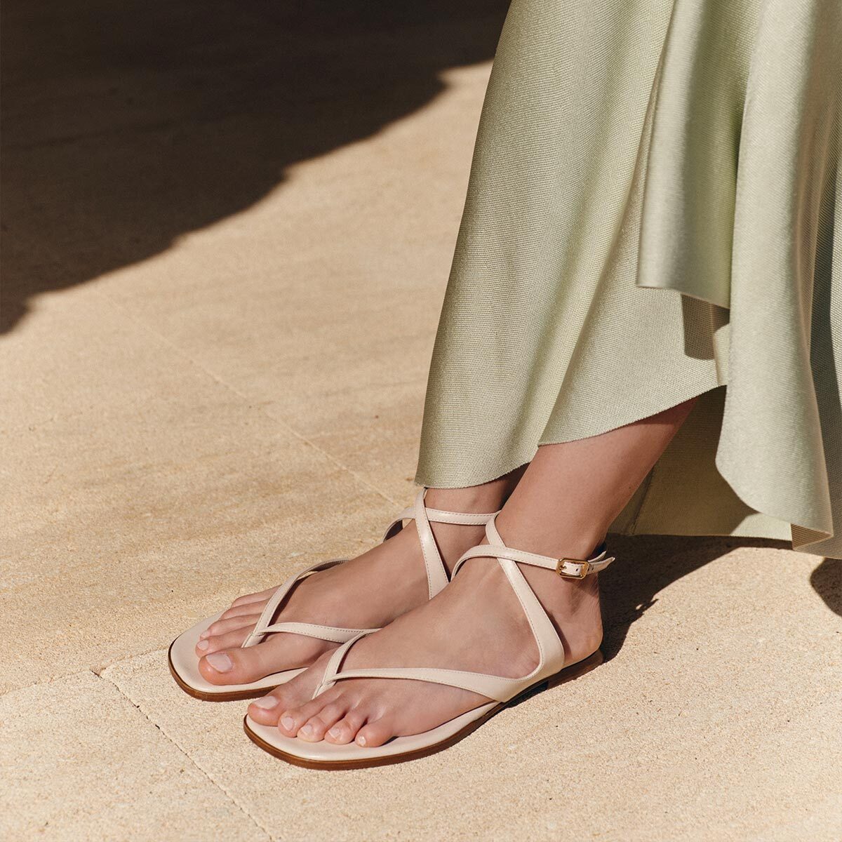A person in Margaux’s The Palermo Sandal - Magnolia Nappa with a cushioned insole and a light green skirt stands on sunlit stone, capturing effortless getaway style.