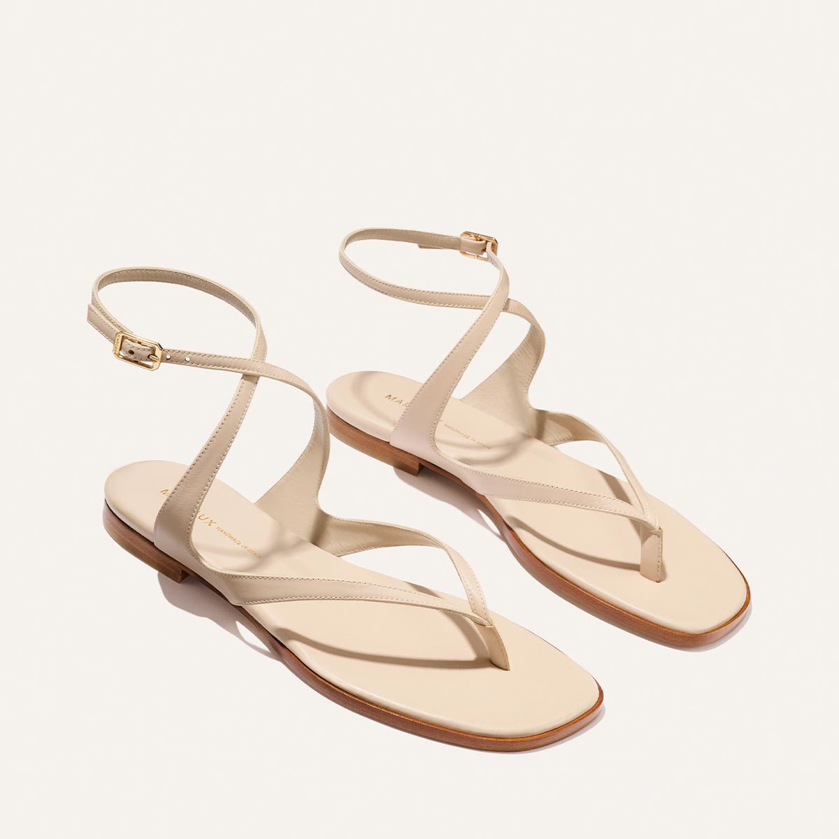 The Palermo Sandal - Magnolia Nappa by Margaux features beige ankle straps with gold buckles, a cushioned insole, minimalist design, and low wooden heels—perfect summer footwear shown on a white background.