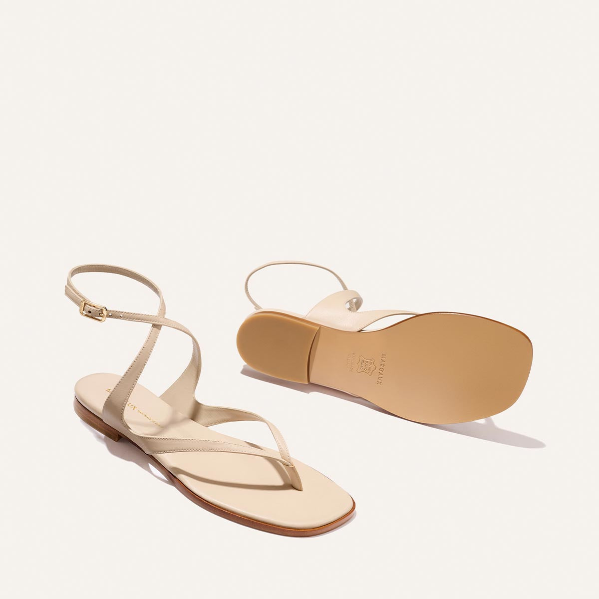 The Palermo Sandal - Magnolia Nappa by Margaux is a stylish pair of beige sandals with ankle straps and flat soles, shown on a white background—one upright, the other flipped to reveal the cushioned insole. Perfect summer footwear.