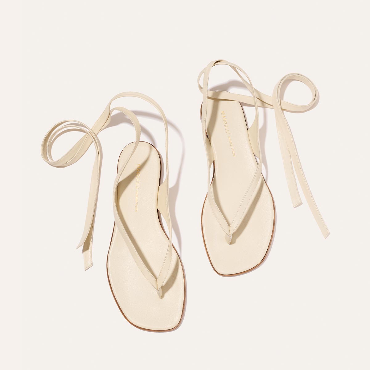 The Margaux Palermo Wrap Sandal in Ivoire Nappa features a minimalist design with square soles and long ankle wrap straps, shown on a plain, off-white background.
