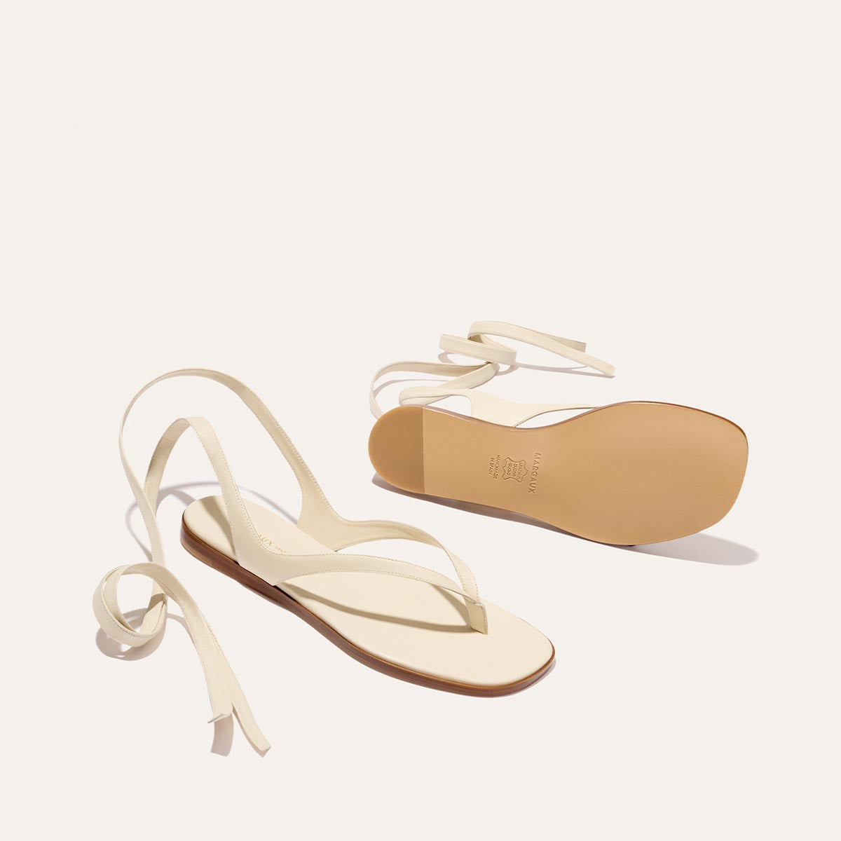 The Margaux Palermo Wrap Sandal in Ivoire Nappa is a minimalist, flat cream sandal with thin ankle wrap straps. One sandal stands upright and the other sole-up, showing the tan sole against a plain light background.
