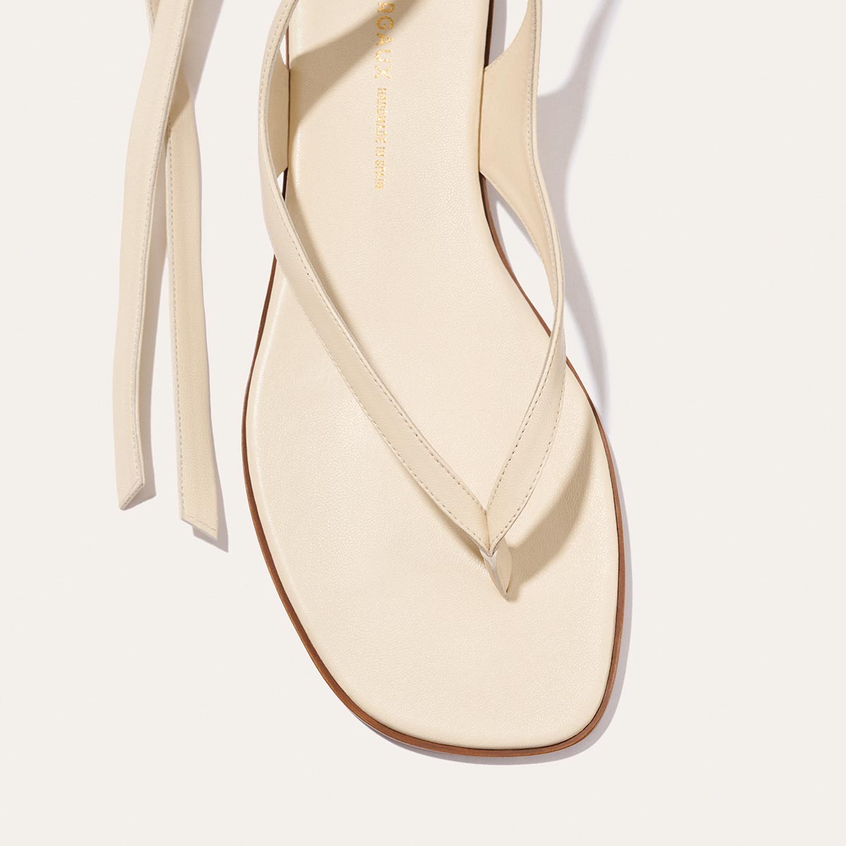 A close-up of Margaux's The Palermo Wrap Sandal in Ivoire Nappa, showcasing its minimalist design with a square toe, thong strap, and long ankle ties, set against a light background.