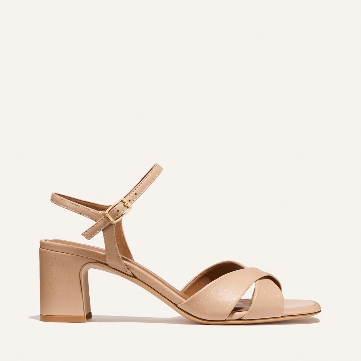 The Paloma Sandal - Dune Nappa by Margaux features a beige chunky heel, single ankle strap with gold buckle, and two crisscross straps over the open toe, displayed against a plain light background.
