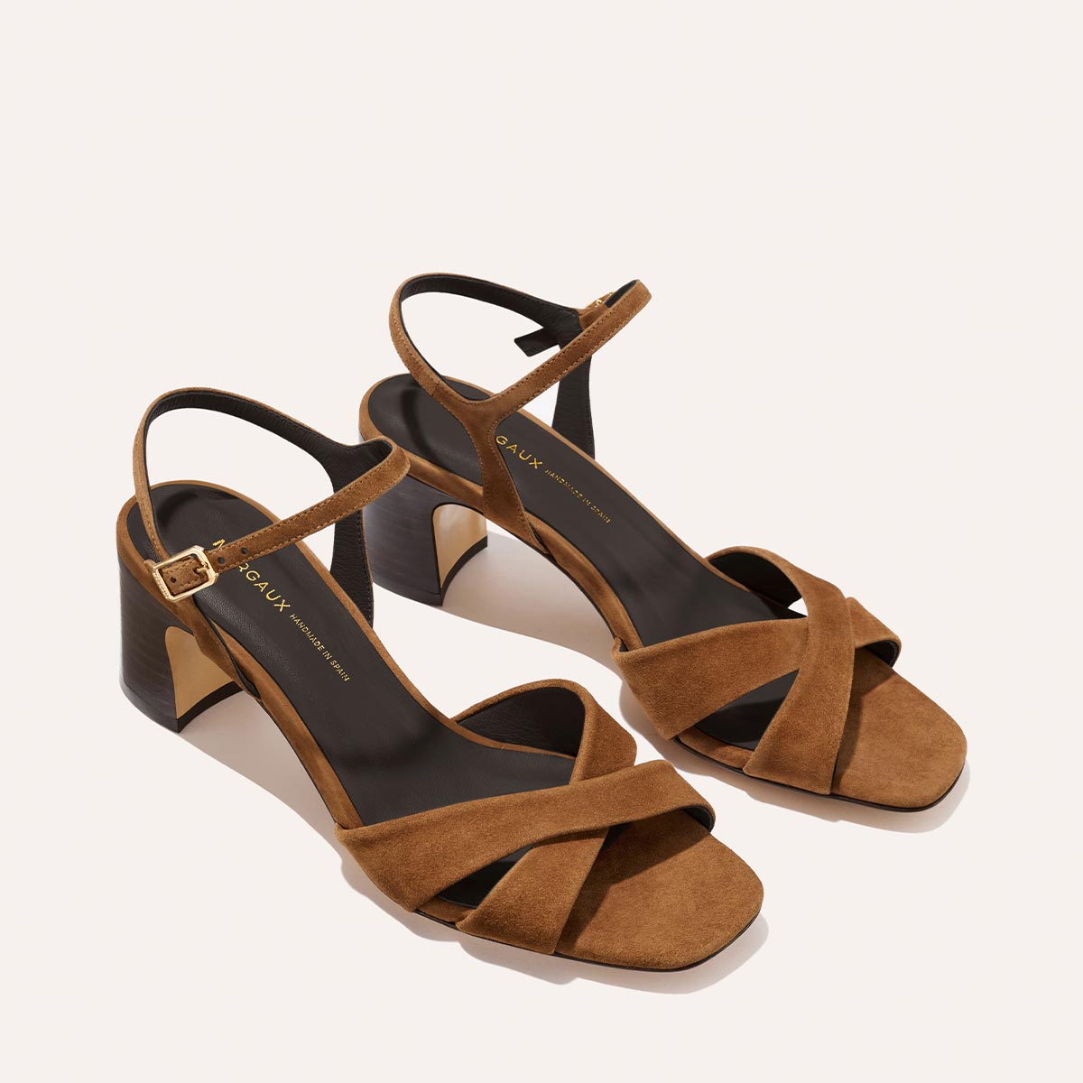 The Paloma Sandal - Nutmeg Suede by Margaux features brown suede, crisscross leather straps, ankle straps with gold buckles, and 2.5-inch dark brown block heels, displayed on a light background.