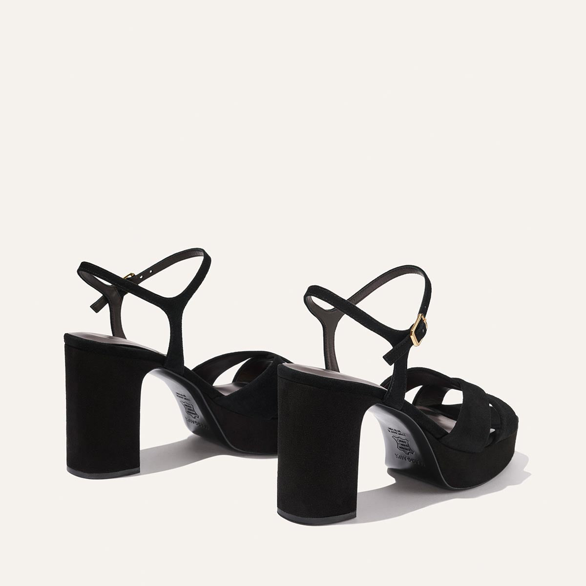 The Paloma Platform by Margaux in black suede features thick block heels, ankle straps with gold buckle closures, and a padded platform for all-night comfort, shown on a light background facing away from the viewer.