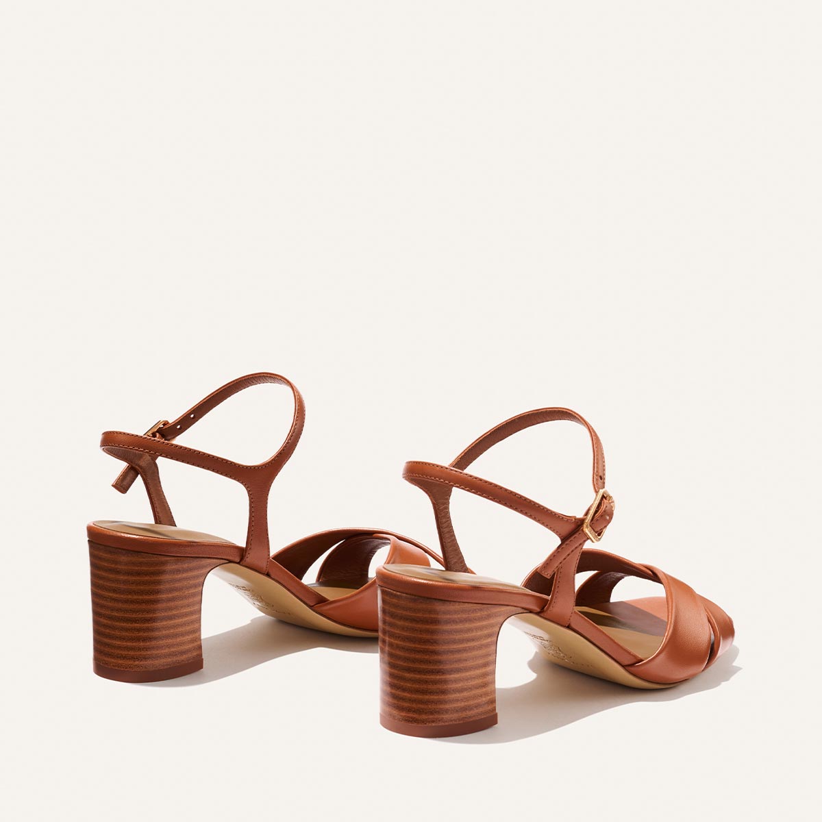 The Paloma Sandal - Saddle Nappa by Margaux features brown leather with crossed toe straps, ankle straps with buckle closures, and thick stacked heels, shown against a light background.