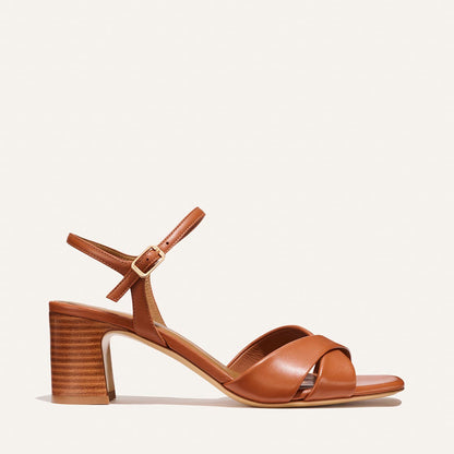 The Paloma Sandal by Margaux in Saddle Nappa features a tan leather finish, block stacked heel, open toe, crisscross front straps, and an ankle strap with a gold buckle. Displayed against a plain light background.