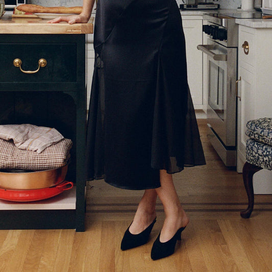 Wearing the Margaux Alison Roman x Margaux: The Parlor Mule in Black Suede, a person stands in a kitchen beside a green drawer and a stack of folded fabrics on a shelf.