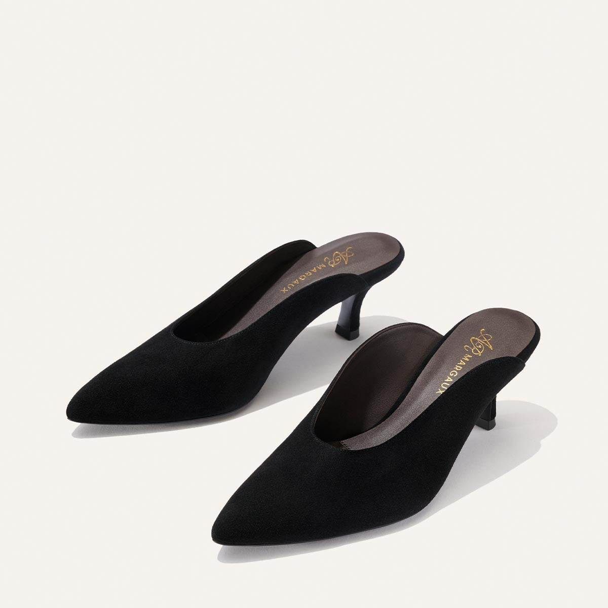 A pair of The Parlor Mule in black suede from Margaux’s Alison Roman collaboration, featuring pointed toes, mid-height kitten heels, and gold Margaux logo text on the insole, displayed against a plain light background.