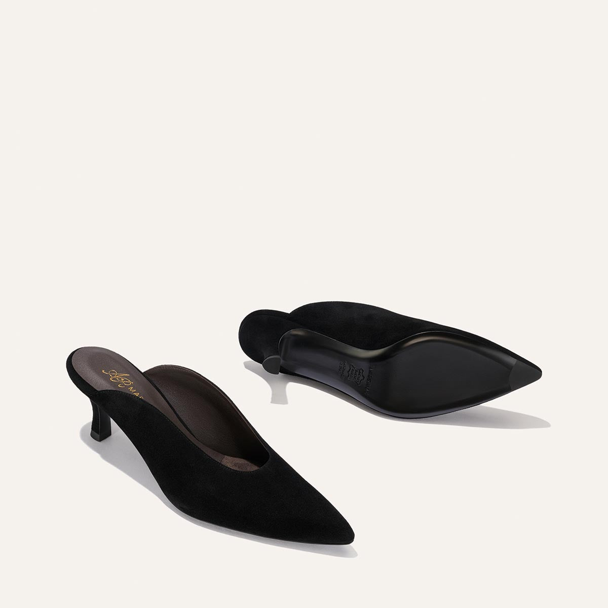 A pair of Margaux Alison Roman x Margaux: The Parlor Mule – Black Suede shoes with pointed toes and low kitten heels. One mule stands upright while the other rests on its side, displaying the sole, set against a light, plain background.