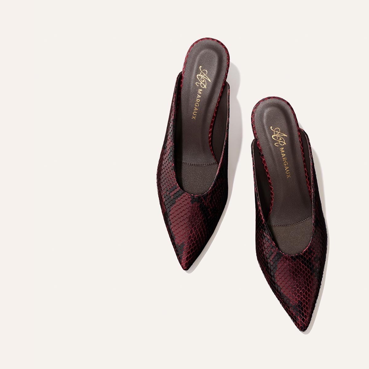 Showcased against a light, off-white background, the Alison Roman x Margaux: The Parlor Mule – Burgundy Python Embossed by Margaux features a pointed toe, slip-on design with low heels and a striking dark red and black snakeskin pattern.