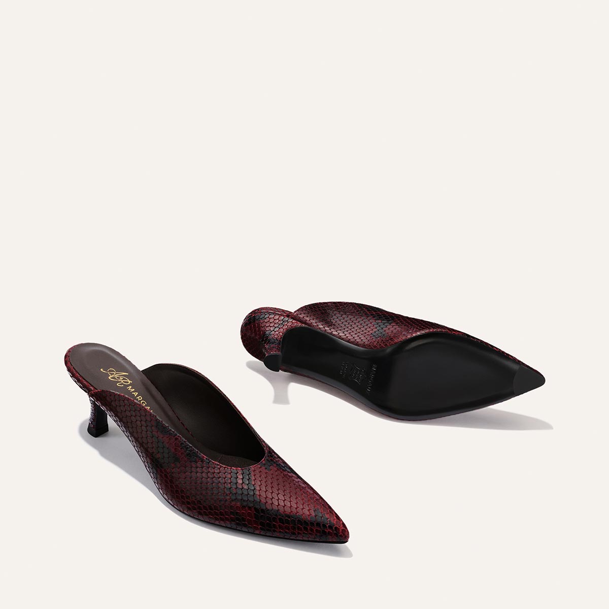A pair of Margaux Alison Roman x Margaux: The Parlor Mule in Burgundy Python Embossed, featuring a snakeskin pattern, pointed toes, and mid-height heels—one upright and one on its side—set against a plain light background.