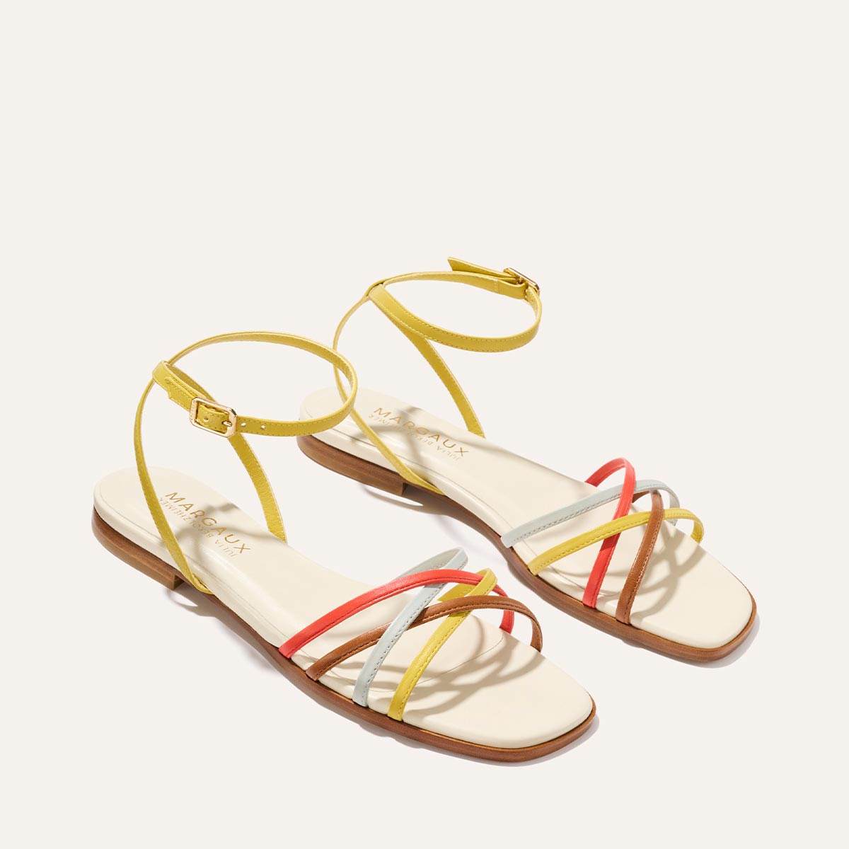 The Paros Sandal by Margaux features thin yellow, silver, and coral straps in cotton and multicolor nappa, ankle buckles, and a light beige footbed—offering a playful look inspired by Julia Berolzheimer's elevated style.