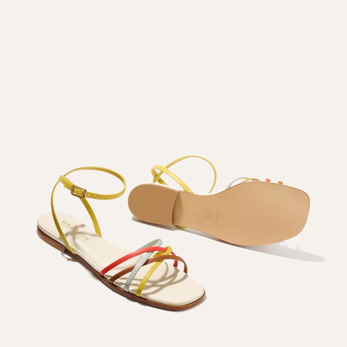 The Margaux Paros Sandal features playful, colorful flat design in cotton and multicolor nappa with thin yellow, orange, and white straps; one sandal shows the sole while the other displays the insole and vibrant straps.