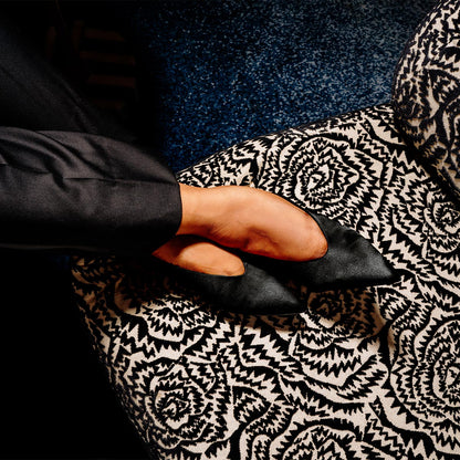 A person in dark clothing wears The Parra - Black Satin shoes by margaux-development, resting their feet on an upholstered chair with a striking black and white geometric floral pattern over a deep blue floor.