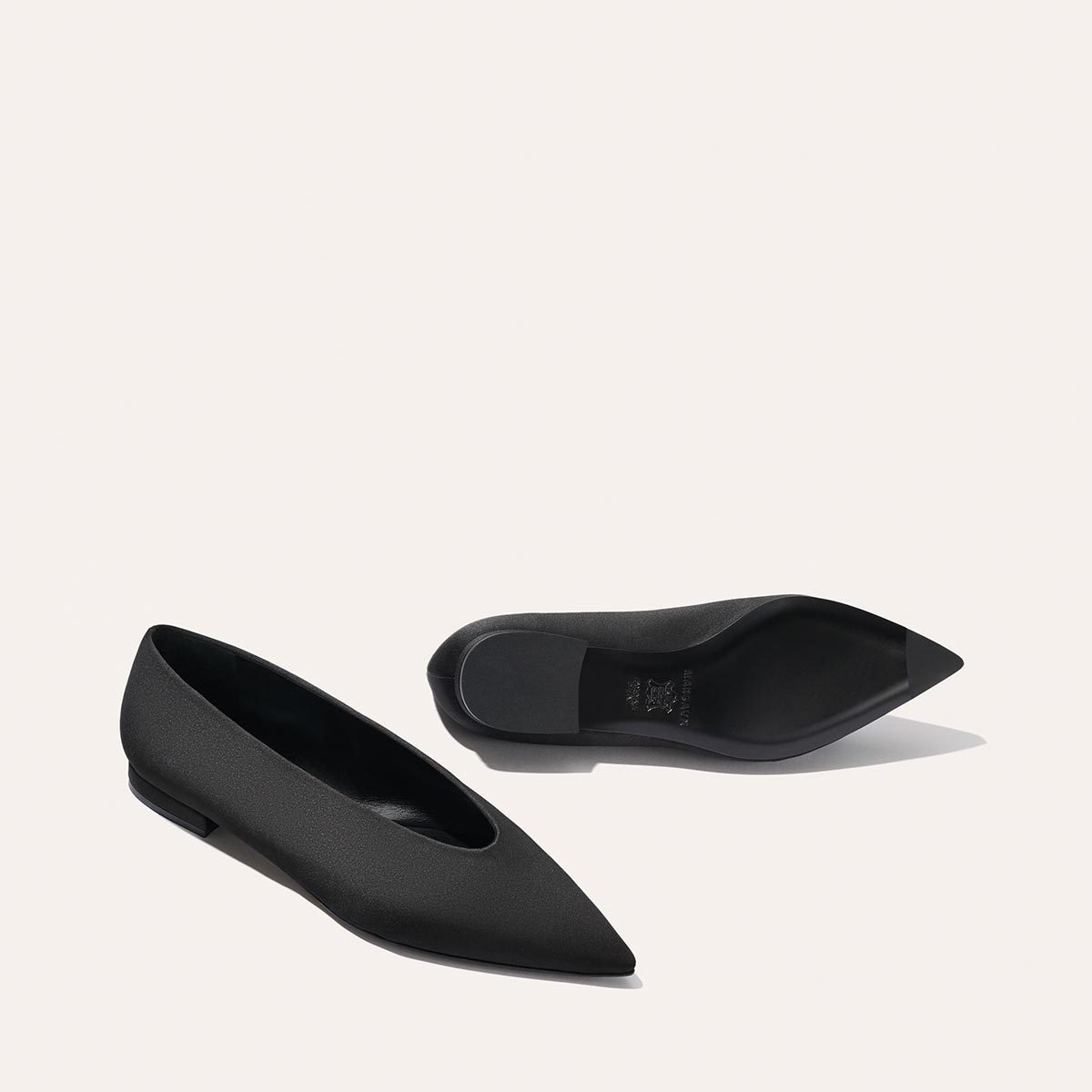 The Parra - Black Satin flats by margaux-development are shown on a light background, with one shoe standing upright and the other lying on its side to reveal the sole.