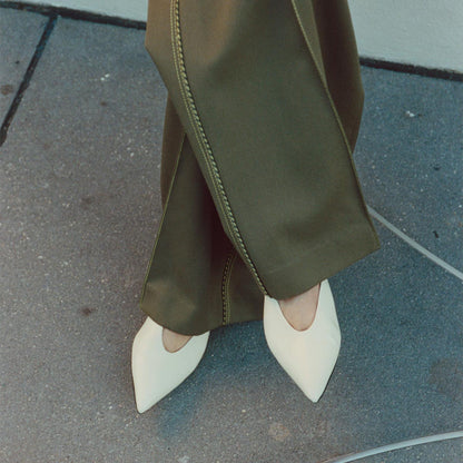 A person stands on a gray sidewalk in olive green pants with prominent stitching, feet crossed to showcase The Parra - Ivoire Nappa pointed-toe flats by margaux-development, which add structure to the outfit’s silhouette.