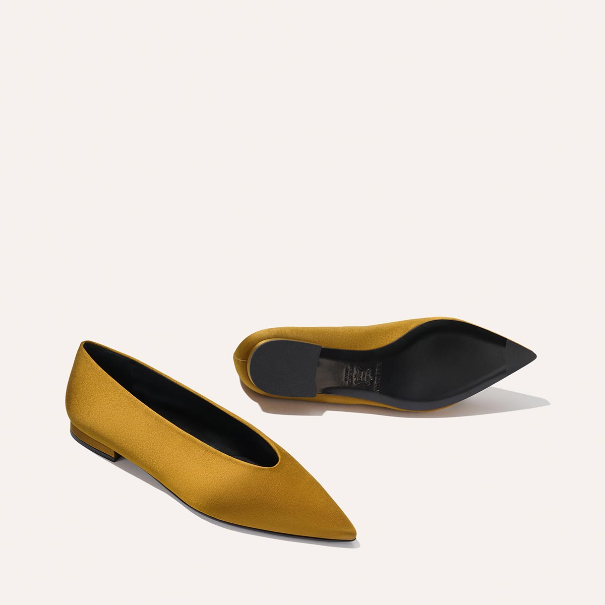 A pair of The Parra - Ochre Satin flats by margaux-development, featuring mustard yellow satin, pointed toes, low heels, and black soles. Displayed on a light background with one upright and the other on its side to show the sole.