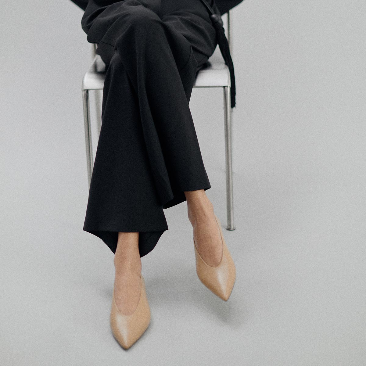 A person sits on a white chair against a light gray background, wearing black pants and The Parra - Praline Nappa flats by margaux-development. Their upper body is not fully visible, drawing attention to the shoes’ structured silhouette.