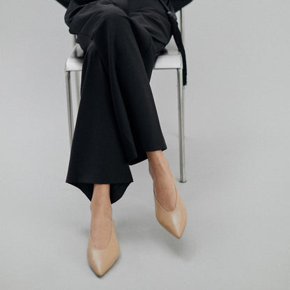 A person sits on a white chair against a light gray background, wearing black pants and The Parra - Praline Nappa flats by margaux-development. Their upper body is not fully visible, drawing attention to the shoes’ structured silhouette.