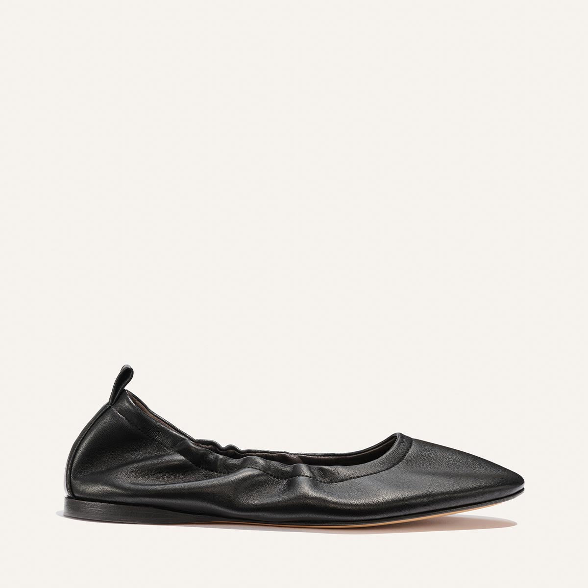 The Margaux "The Pascale - Black Nappa" is a black leather ballet flat with a pointed toe, elasticized ruffled edge, and small heel pull tab, displayed against a plain light background.