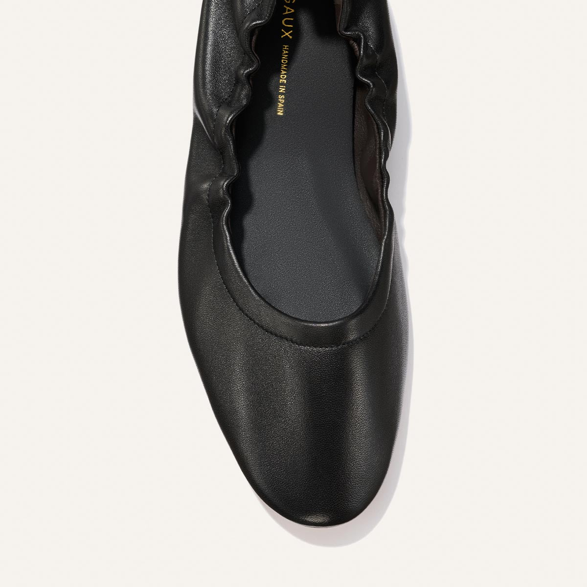 A top view of Margaux's The Pascale - Black Nappa flat shoe, featuring black leather with a ruched edge along the opening and a rounded toe, shown on a plain white background.