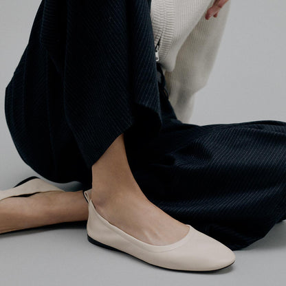 A person kneels on the floor wearing black pinstripe pants, a cream sweater, and Margaux’s The Pascale - Bone Nappa ballet flats, featuring an elasticated back and slight heel strap, against a plain grey background.