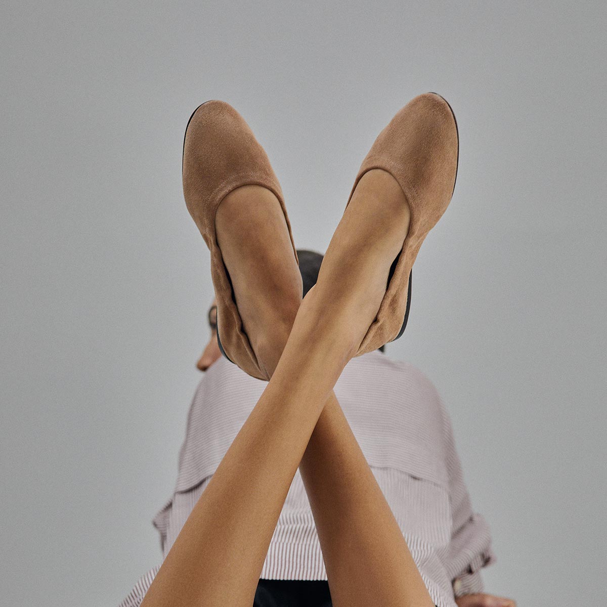A person with crossed legs wears Margaux's The Pascale - Cameo Suede beige ballet flats with an elasticated back, seen from below against a simple gray background.