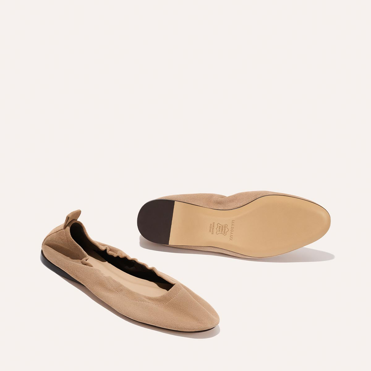 A pair of Margaux’s The Pascale - Cameo Suede ballet flats in beige, featuring a ruched edge and back pull tab—one on its side, the other sole-up—displayed on a light neutral background.