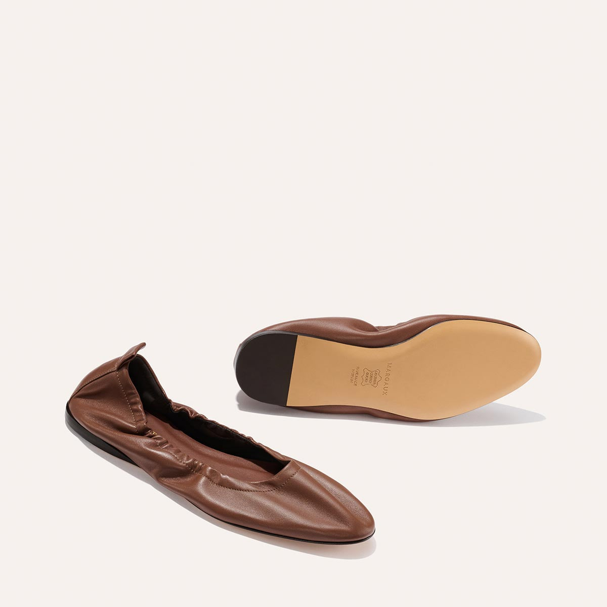 A pair of Margaux "The Pascale - Cognac Nappa" ballet flats: one upright to show the brown leather upper, the other flipped to display the tan sole, both positioned on a plain light background.